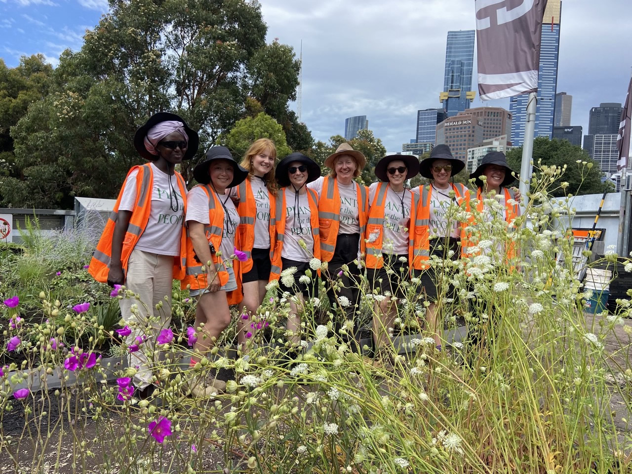 Group of women gardeners wearing sun hats and orange high vis PPE standing on a roof garden. There are flowers in the foreground and trees and the tops of highrise buildings in the background.