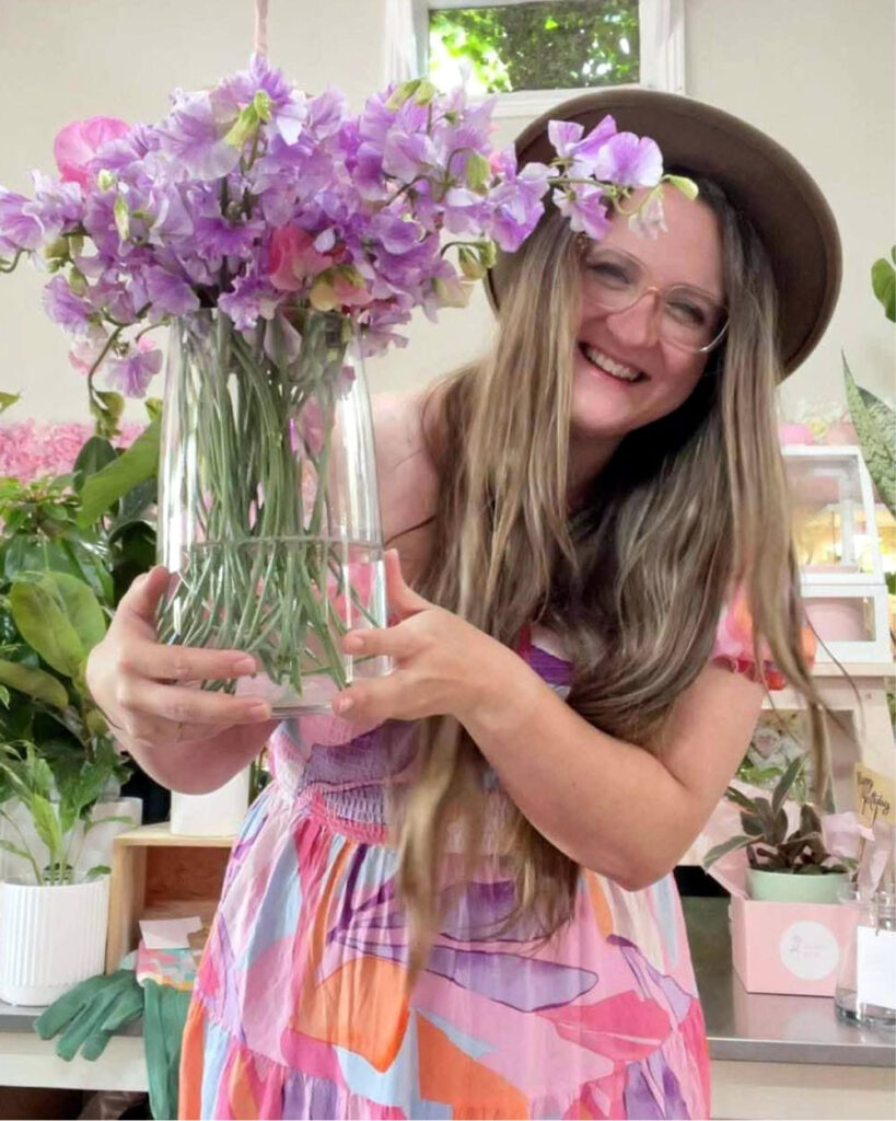 Photo of Sarah Vafidis wearing a pastel coloured dress, hat, and holding a vase of lilac coloured pea flowers.