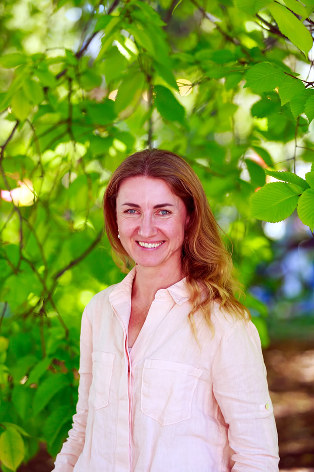 Photo of Magdalena Kazmierczak. She has long brown hair and is wearing a light coloured shirt. She is standing in front of a green tree.