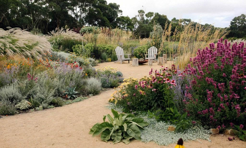Photo of a garden with pretty floral borders and couple of white chairs along a path.
