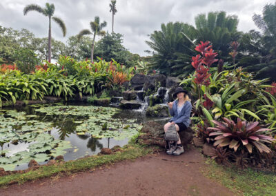 Photo of Liesha Northover wearing a hat and hiking boots sitting on a rock in gardens at the Dole Plantation. There is large pond covered in lily pads, palm trees and other tropical plants around her.
