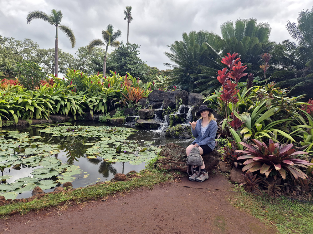 Photo of Liesha Northover wearing a hat and hiking boots sitting on a rock in gardens at the Dole Plantation. There is large pond covered in lily pads, palm trees and other tropical plants around her.