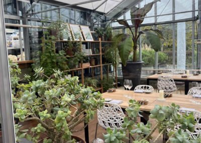 Inside the cafe at Restaurant De Kas. There are tables, chairs, bookshelves with plants and items on them, large pot plants around, and the restaurant looks like a glasshouse.